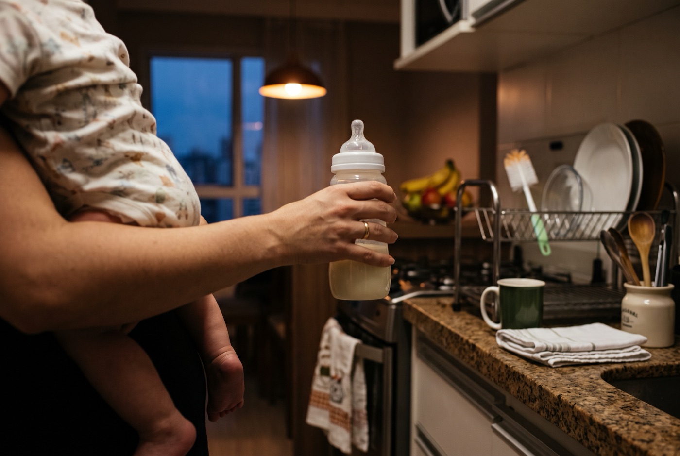 Mão de mãe segurando a mamadeira enquanto o outro braço carrega o bebê, na cozinha, no fim do dia.